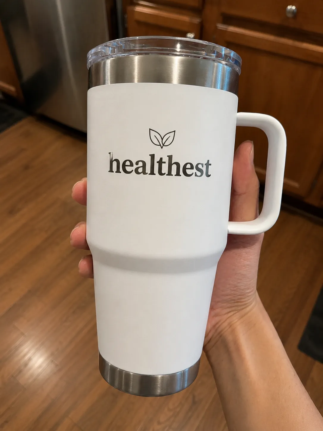 A hand holding the HEALTHEST 30 oz Insulated Tumbler, a white double-wall stainless-steel tumbler with a clear flip lid, comfort handle, and the engraved HEALTHEST leaf logo.